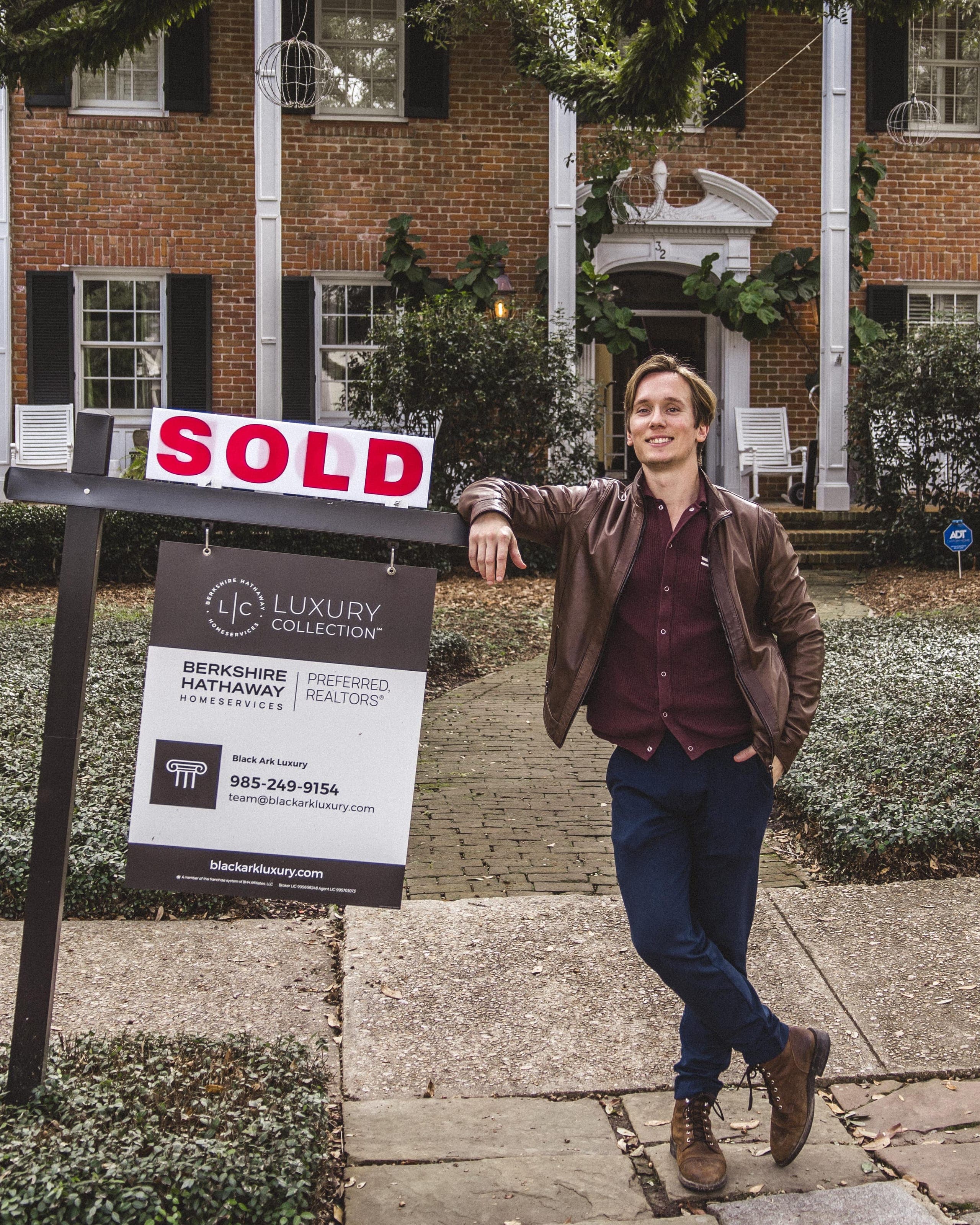 Garrett Shearman leaning on a SOLD sign in front of a two-story brick colonial home